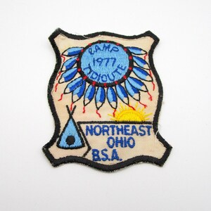 May include: Embroidered patch with a cream-colored background and a black border. The patch features a blue circle with the words "CAMP HIDIOUTE 1977" and blue feather designs. Below is a yellow sun, a blue teepee, and the words "NORTHEAST OHIO B.S.A."