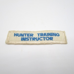May include: A rectangular patch with the words "HUNTER TRAINING INSTRUCTOR" embroidered in blue on an off-white background. The patch has a stitched border and a slightly frayed edge.