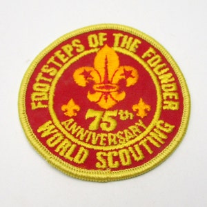 May include: A round, red and gold patch with the text "FOOTSTEPS OF THE FOUNDER" and "75th ANNIVERSARY WORLD SCOUTING". The patch features a yellow fleur-de-lis symbol and a gold border.