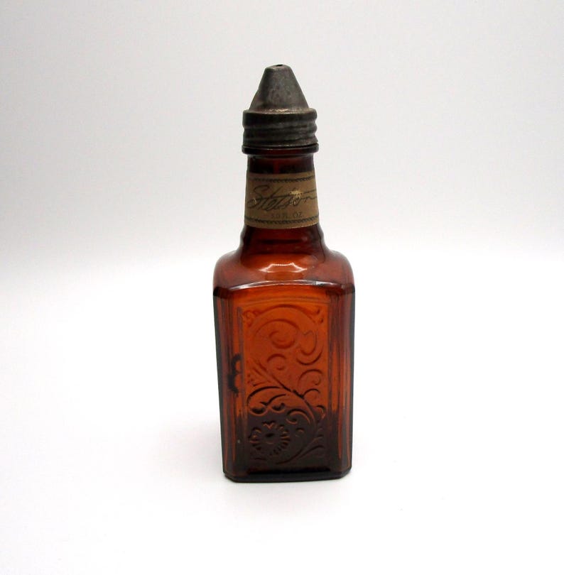 Puede incluir: Una botella vintage de vidrio &aacute;mbar con un tap&oacute;n de metal. La botella tiene un dise&ntilde;o floral en relieve y una etiqueta que dice "Stato" en cursiva.
