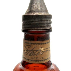 Puede incluir: Primer plano de una botella de vidrio marr&oacute;n con una tapa de metal. La botella tiene una etiqueta que dice "Stetson" y "5.0 FL. OZ."