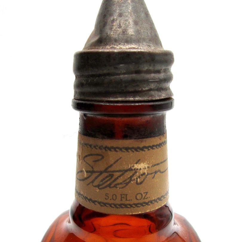 Puede incluir: Primer plano de una botella de vidrio marr&oacute;n con una tapa de metal. La botella tiene una etiqueta que dice "Stetson" y "5.0 FL. OZ."