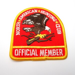 May include: Embroidered patch with a red background and a yellow border. The patch features a brown and white eagle perched on a rifle, with the text "NORTH AMERICAN HUNTING CLUB" and "OFFICIAL MEMBER" in white.