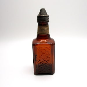 Puede incluir: Una peque&ntilde;a botella de vidrio &aacute;mbar con un tap&oacute;n de metal. La botella tiene una etiqueta que dice "Sharon" y est&aacute; grabada con un dise&ntilde;o floral.