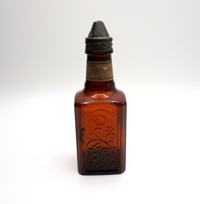 Puede incluir: Una peque&ntilde;a botella de vidrio &aacute;mbar con un tap&oacute;n de metal. La botella tiene una etiqueta que dice "Sharon" y est&aacute; grabada con un dise&ntilde;o floral.