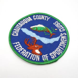 May include: Embroidered patch with a blue background, featuring a green and white fish, a brown deer, and the text "Chautauqua County Federation of Sportsmen's Clubs" in white. The patch has a green border.
