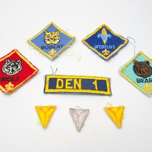 May include: A collection of Boy Scout patches, including Wolf, Bobcat, Bear, Webelos, and a Den 1 patch. There are also three yellow triangular patches.