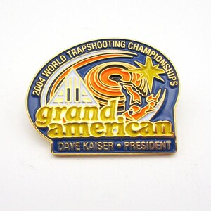 May include: A blue, gold, and orange enamel pin from the 2004 World Trapshooting Championships. The pin features the words "grand american" in gold lettering, along with the name "Dave Kaiser - President."