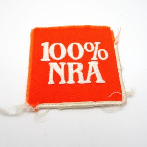 May include: Square orange patch with white embroidered text reading "100% NRA". The patch has a white border and frayed edges. The background is plain white.