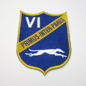May include: A blue shield-shaped patch with a yellow border. The patch features the Roman numeral VI in white, a white running dog, and the text "PRIMUS-INTER-PARES" in yellow.