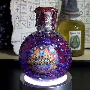Op de afbeelding: Een ronde, paarse glazen fles gevuld met glitter en vloeistof, met het label "Unicorn Droppings". De fles heeft een zilveren hals met druipend rood was en staat op een zilveren, oplichtende basis. Op de achtergrond staat een tweede fles met een heksenhoedvormige stopper.
