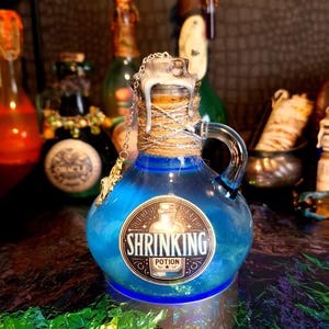 May include: A blue glass potion bottle with a handle, labeled "SHRINKING POTION." The bottle has a cork stopper wrapped with twine and a silver chain. Other bottles are in the background. The bottle sits on a silver base.