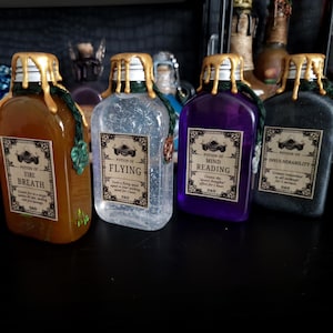 Puede incluir: Cuatro botellas de poción decorativas con sellos de cera dorados y etiquetas. Las botellas están llenas de líquidos de diferentes colores: marrón, transparente, morado y negro. Cada etiqueta dice "Poción de" seguido de un efecto diferente.