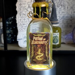 Op de afbeelding: Een heldere glazen fles met gele vloeistof, met het opschrift "Potion of Electricity". De fles heeft een bruine kurk, een zwarte hals en gouden draaddetails. Hij staat op een zilveren, oplichtende basis. De achtergrond toont alchemistische symbolen.