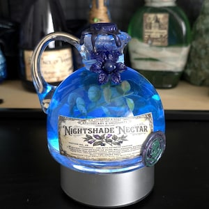 Può includere: Una boccetta rotonda in vetro blu con manico, riempita di liquido e vegetazione. Presenta un'etichetta con la scritta Nightshade Nectar, un sigillo in cera viola scuro con un teschio e un fiore viola sul collo, poggiata su una base argentata.