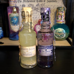 Op de afbeelding: Twee decoratieve glazen flessen met waszegels. Eén fles is gevuld met een lichtgele vloeistof en is gelabeld met "Supernova Solstice - The Light". De andere fles bevat een donkerpaarse vloeistof en is gelabeld met "Umbral Eclipse - The Dark".