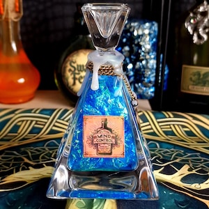 Op de afbeelding: Een heldere, driehoekige glazen fles gevuld met blauwe vloeistof en een label met de tekst "Potion Mind Control". De fles heeft een heldere, diamantvormige stop en is versierd met een gouden ketting. De fles staat op een patroon oppervlak.