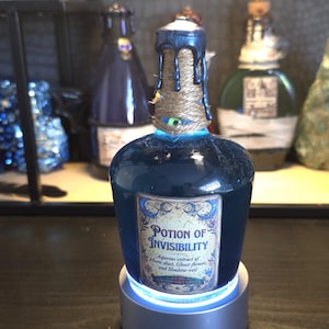 May include: A glass bottle filled with blue liquid, labeled Potion of Invisibility, Aqueous extract of Moon-dust, Ghost-flower, and Shadow-veil. The neck is wrapped in twine with dark blue wax drips and a small green eye, resting on a light base.