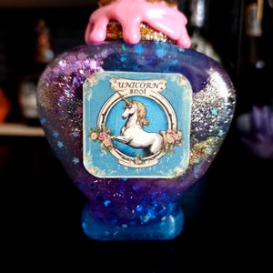 May include: A heart-shaped bottle filled with blue glitter, labelled "Unicorn Snot." The bottle has a pink, dripped-style top and a gold glitter base. The label features a white unicorn. The bottle is approximately 10 cm tall.