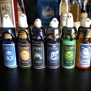 Op de afbeelding: Zes miniatuur glazen flesjes met kurken stoppen bekroond met witte spookfiguurtjes. Elk flesje heeft een etiket met een unieke naam: Chilly Essence, Glowy Serum, Spooky Potion, Gloomy Extract, Slimy Brew en Floaty Elixir, met zwarte wasdruppels.