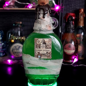 Puede incluir: Una botella de vidrio verde con la etiqueta "Mummy Extract" y un tapón de corcho coronado con una calavera. La botella está envuelta en tela blanca y tiene un asa. Se encuentra sobre una base verde brillante, con otras botellas al fondo.
