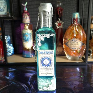 Può includere: Una bottiglia di vetro rettangolare riempita di liquido blu, con un tappo sigillato con cera argentata, decorazioni in filigrana argentata agli angoli e un'etichetta con la scritta Moon Water Celestial Elixir, con una catenella argentata al collo.