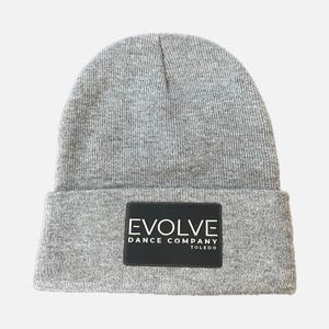 May include: A gray knit beanie with a folded brim. A black rectangular patch on the front displays the white text "EVOLVE DANCE COMPANY TOLEDO". The beanie is made of a soft, textured material.