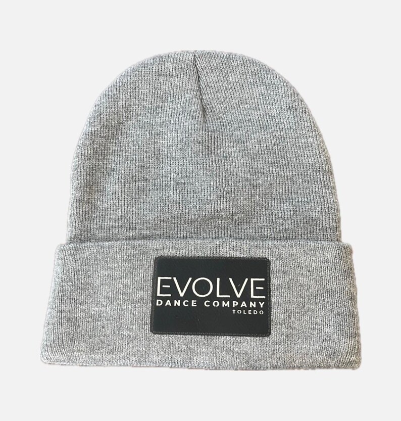 May include: A gray knit beanie with a folded brim. A black rectangular patch on the front displays the white text "EVOLVE DANCE COMPANY TOLEDO". The beanie is made of a soft, textured material.