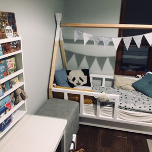 May include: A white wooden bed frame with a teepee-style canopy, featuring a white and gray patterned bedding, a blue and white patterned pillow, and a black and white panda pillow. A white bookshelf with a variety of children's books is visible in the background.
