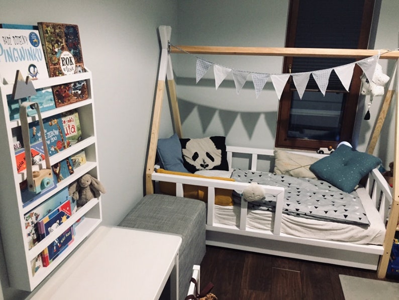 May include: A white wooden bed frame with a teepee-style canopy, featuring a white and gray patterned bedding, a blue and white patterned pillow, and a black and white panda pillow. A white bookshelf with a variety of children's books is visible in the background.