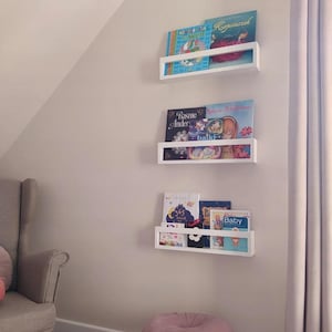 May include: Three white floating bookshelves mounted on a light-colored wall, each holding colorful children's books. A gray armchair and a pink ottoman are in the foreground. The books have titles in different languages.
