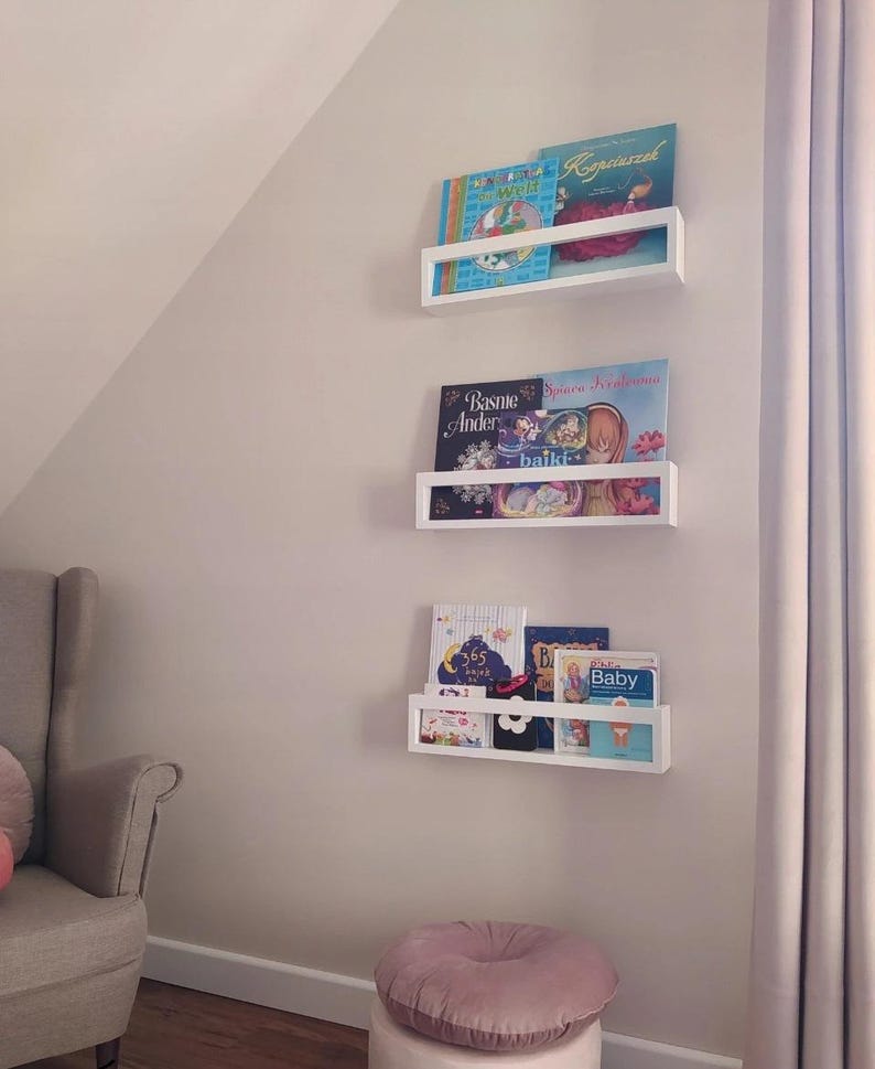 May include: Three white floating bookshelves mounted on a light-colored wall, each holding colorful children's books. A gray armchair and a pink ottoman are in the foreground. The books have titles in different languages.