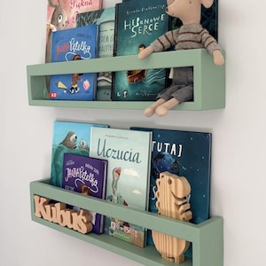 May include: Sage green floating bookshelves filled with children's books. A stuffed mouse sits on the top shelf. The bottom shelf has a wooden name cutout that says "Kubuś".