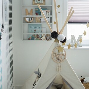 May include: A white teepee tent with a gray base and a wooden sign that says "Dream Big" hanging from the top. The teepee is decorated with gold stars and a white and black pom-pom garland. The teepee is set up in a room with a white wall and a bookshelf with books and toys.