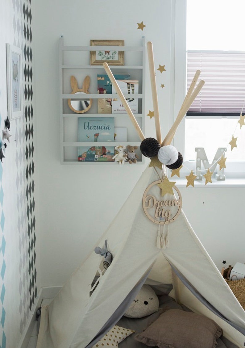 May include: A white teepee tent with a gray base and a wooden sign that says "Dream Big" hanging from the top. The teepee is decorated with gold stars and a white and black pom-pom garland. The teepee is set up in a room with a white wall and a bookshelf with books and toys.