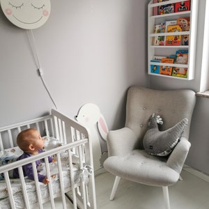 May include: A white crib with a baby in it, a white armchair with a gray pillow, a white bookshelf with colorful children's books, and a white wall with a smiling moon wall light.