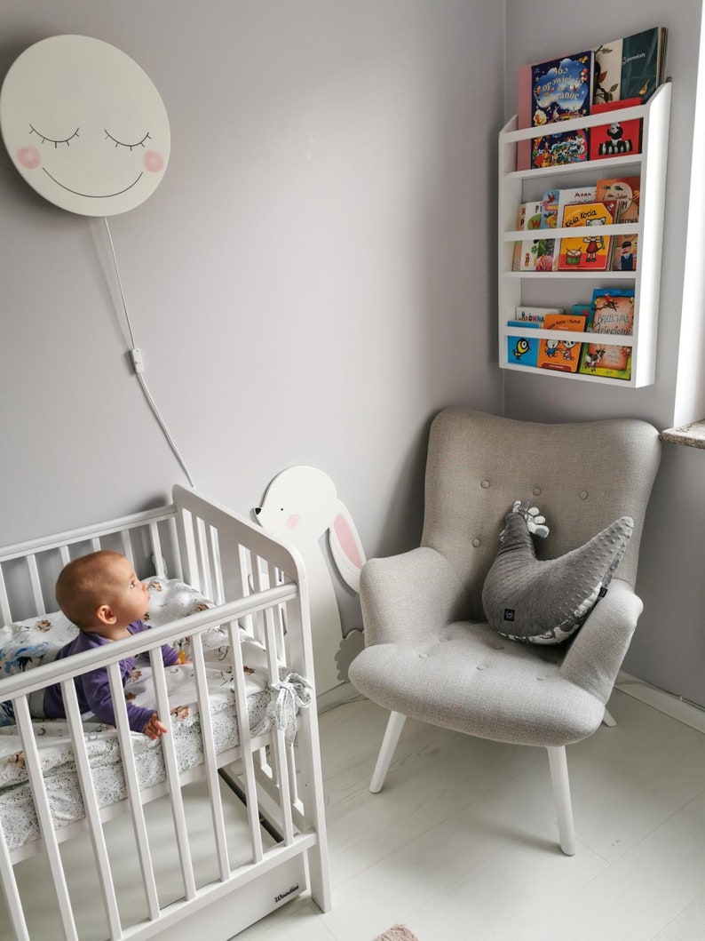 May include: A white crib with a baby in it, a white armchair with a gray pillow, a white bookshelf with colorful children's books, and a white wall with a smiling moon wall light.