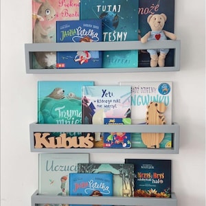 May include: Three gray floating shelves filled with children's books. The books have colorful covers with illustrations and text. A small stuffed bear and wooden dinosaur are on the shelves. The word "Kubus" is displayed on the middle shelf.