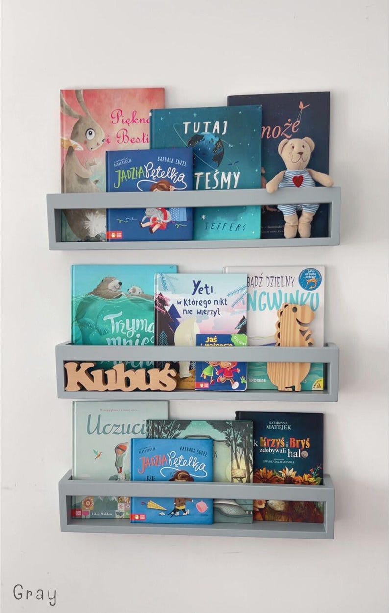 May include: Three gray floating shelves filled with children's books. The books have colorful covers with illustrations and text. A small stuffed bear and wooden dinosaur are on the shelves. The word "Kubus" is displayed on the middle shelf.