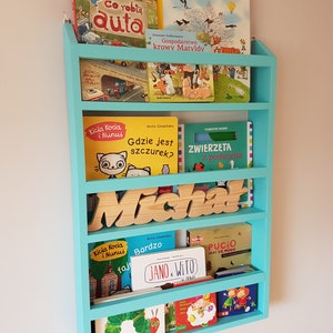 May include: A light blue bookshelf with five shelves, filled with children's books. The top shelf has a book titled "Wiosna na ulicy Czereśniowej" and another titled "Gospodarstwo krow Matyldy". The second shelf has a book titled "Kicia Kocia i Nunus". The third shelf has a book titled "Gdzie jest szczurek?" and another titled "Zwierzęta z podw&oacute;rka". The fourth shelf has a wooden name sign that says "Michał". The fifth shelf has a book titled "Kicia Kocia i Nunus" and another titled "Jano. Wito". The bottom shelf has a book titled "Pucio uczy się m&oacute;wić".