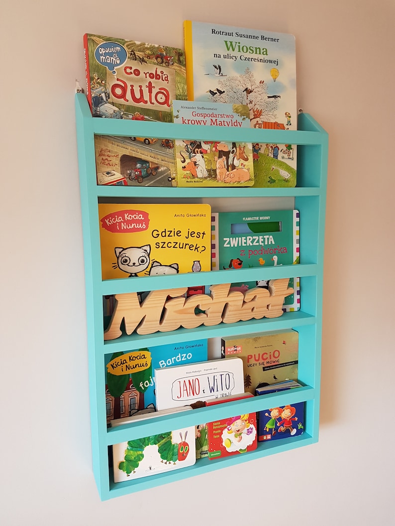 May include: A light blue bookshelf with five shelves, filled with children's books. The top shelf has a book titled "Wiosna na ulicy Czereśniowej" and another titled "Gospodarstwo krow Matyldy". The second shelf has a book titled "Kicia Kocia i Nunus". The third shelf has a book titled "Gdzie jest szczurek?" and another titled "Zwierzęta z podw&oacute;rka". The fourth shelf has a wooden name sign that says "Michał". The fifth shelf has a book titled "Kicia Kocia i Nunus" and another titled "Jano. Wito". The bottom shelf has a book titled "Pucio uczy się m&oacute;wić".