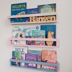 May include: Three wall-mounted bookshelves in light gray, light pink, and gray blue, each holding children's books. The top shelf has the name "Hansen" in wooden letters. The shelves are filled with colorful book covers.