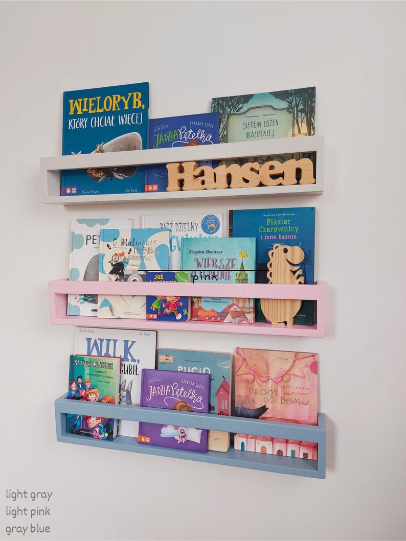 May include: Three wall-mounted bookshelves in light gray, light pink, and gray blue, each holding children's books. The top shelf has the name "Hansen" in wooden letters. The shelves are filled with colorful book covers.