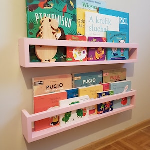 May include: Pink wooden wall-mounted bookshelves filled with colorful children's books. A wooden dinosaur-shaped decoration sits on the top shelf. The shelves are mounted on a light gray wall.