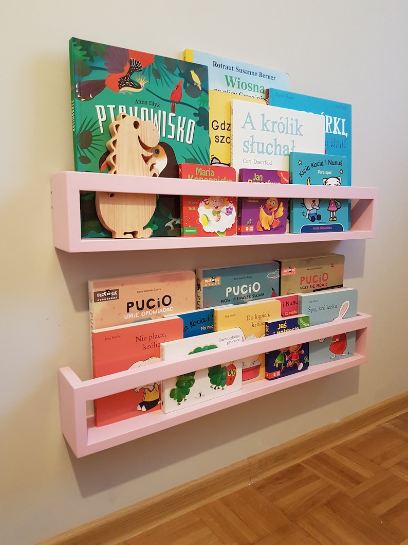 May include: Pink wooden wall-mounted bookshelves filled with colorful children's books. A wooden dinosaur-shaped decoration sits on the top shelf. The shelves are mounted on a light gray wall.