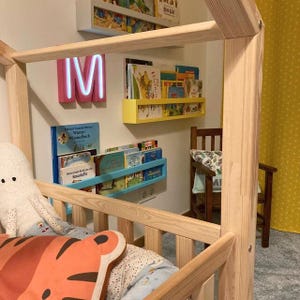 May include: A wooden bed frame with a house-like structure. The bed is decorated with a plush octopus and a tiger-shaped pillow. Bookshelves in yellow, blue, and white hold various books. A pink neon "M" sign is mounted on the wall.
