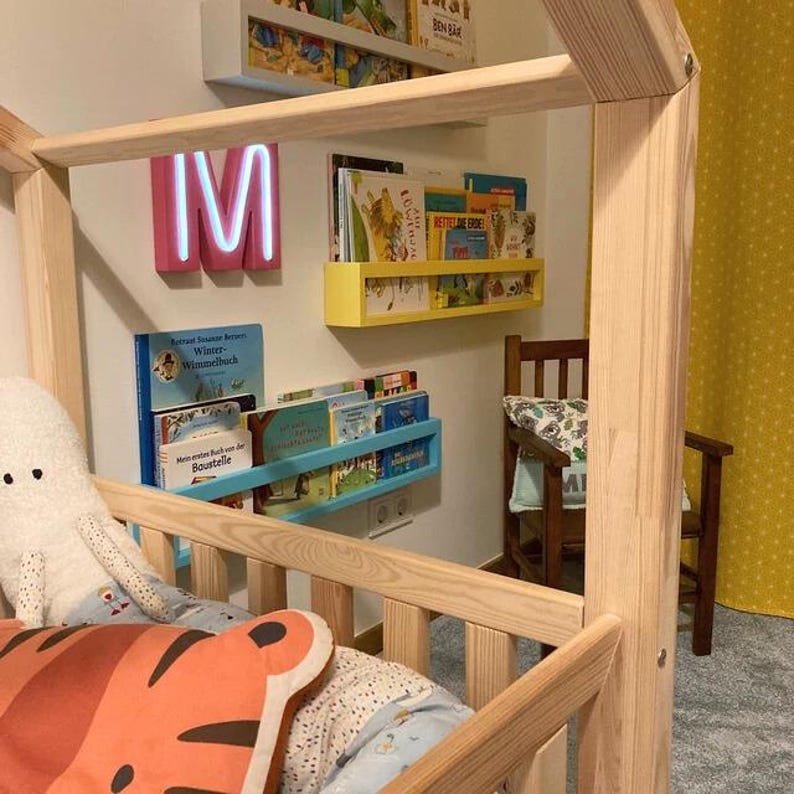 May include: A wooden bed frame with a house-like structure. The bed is decorated with a plush octopus and a tiger-shaped pillow. Bookshelves in yellow, blue, and white hold various books. A pink neon "M" sign is mounted on the wall.