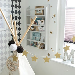 May include: A white teepee tent with a wooden sign that says "Dream Big" hanging from the top. The teepee is decorated with a string of gold star garland and white and brown pom-poms. A white bookshelf with books and a stuffed animal is on the wall behind the teepee.