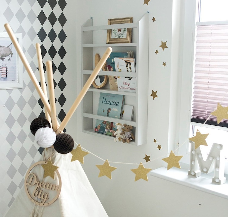 May include: A white teepee tent with a wooden sign that says "Dream Big" hanging from the top. The teepee is decorated with a string of gold star garland and white and brown pom-poms. A white bookshelf with books and a stuffed animal is on the wall behind the teepee.