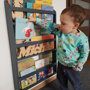 May include: A dark gray wooden bookshelf with multiple shelves filled with children's books. The bookshelf is mounted to a white wall. A young child is standing in front of the bookshelf, reaching for a book. The bookshelf has the name "Micha" spelled out in wooden letters.